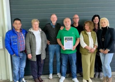 Eine Gruppe von Leuten, die vor einem Gebäude steht. Eine Person hält eine Urkunde in der Hand.