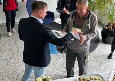 Ein Mann in einem schwarzen Sako der einem Herren, der als Hausmeister tätig ist, eine Urkunde übergibt.