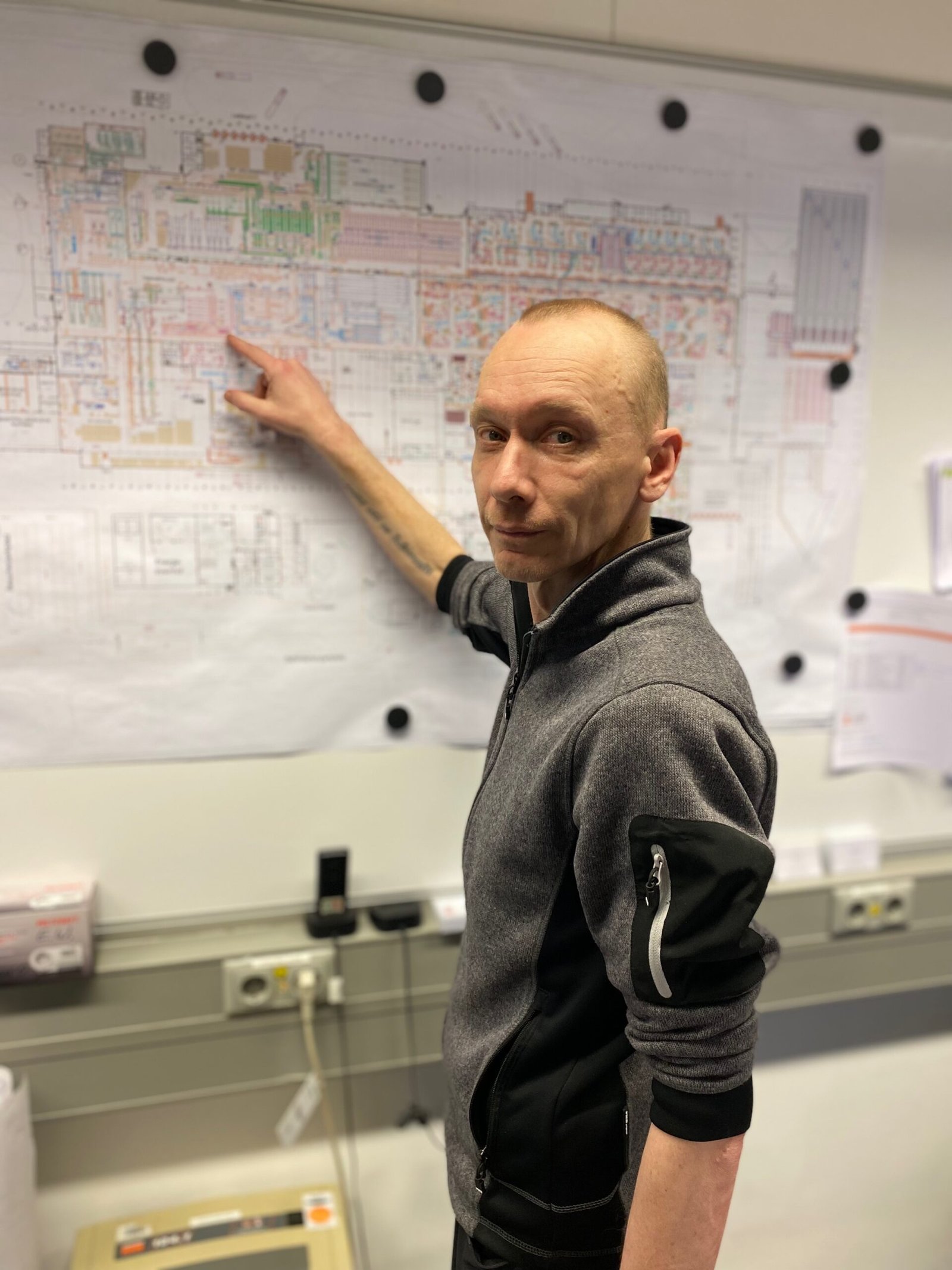 Ein Mitarbeiter aus dem Facility Management, der in die Kamera schaut und mit einem Finger auf einen Plan von einem großen Werksgelände zeigt.