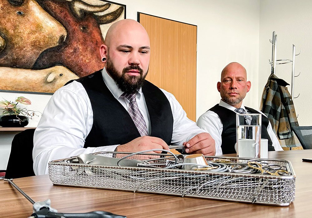 Zwei Herren, die an einem Meetingtisch sitzen und ein Sieb mit Sterilgut vor sich haben. Einer der Herren hält eine Zange des Sterilguts in der Hand und erklärt diese.