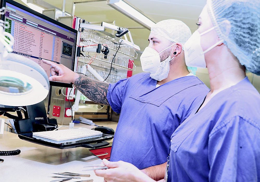 Ein junger Mann und eine junge Dame in Dienstkleidung in einer Sterilgutversorgung im Krankenhaus. Sie besprechen gerade etwas vor einem Computer und halten sauberes Sterilgut in der Hand.