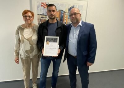 Eine Frau, ein Mann, der eine Urkunde in der Hand hält und ein weiterer Mann.